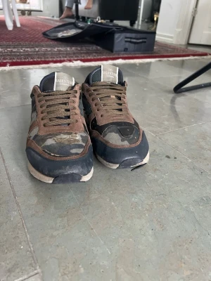 Valentino Garavani camo sneakers - Snygga sneakers från Valentino Garavani med camomönster i grönt, brunt och svart. Skorna har bruna mockadetaljer, gröna skosnören och en vit logga på plösen. Yttermaterialet är en mix av mocka och textil för en cool streetwear-look.