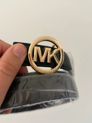 Svart bälte från Michael Kors - Helt nytt svart bälte från Michael Kors med rund, guldig spänne där MK-loggan syns tydligt. Bältet är i slätt skinn och passar perfekt till jeans eller byxor för en trendig look. 110 cm. Jättesnyggt o ha till en jeanskjol i sommar 😍