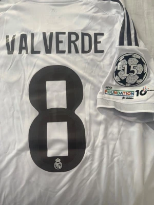 Real Madrid Valverde matchtröja M - Snygg vit Real Madrid matchtröja från Adidas med Valverde och nummer 8 på ryggen. Slim fit-modell med svarta detaljer och officiella UEFA Champions League-märken på ärmen. Perfekt för dig som älskar fotboll och vill bära ditt favoritlag.