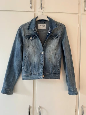 Blå jeansjacka från One Love - Snygg blå jeansjacka från One Love med klassisk krage, silverfärgade knappar och två bröstfickor med lock. Jackan har en tvättad look och raka ärmar. Perfekt att slänga över en hoodie eller t-shirt för en avslappnad stil.
