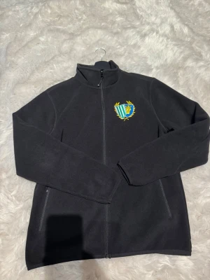 Svart fleece från STHLM SÖDRA *Oanvänd* - Mjuk svart fleecejacka från South West med hel dragkedja och ståkrage. På bröstet finns ett broderat klubbmärke i grönt, blått och gult. Jackan har två sidofickor med dragkedja och är perfekt för kyliga dagar.