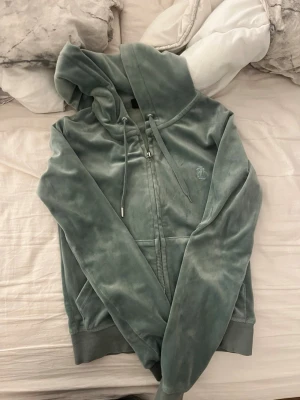 Grön velour hoodie från Juicy Couture - Mjuk och skön hoodie i grön velour från Juicy Couture. Dragkedja framtill, fickor och justerbar huva med snören. Diskret broderad JC-logga på bröstet. Perfekt för en chill och avslappnad stil.