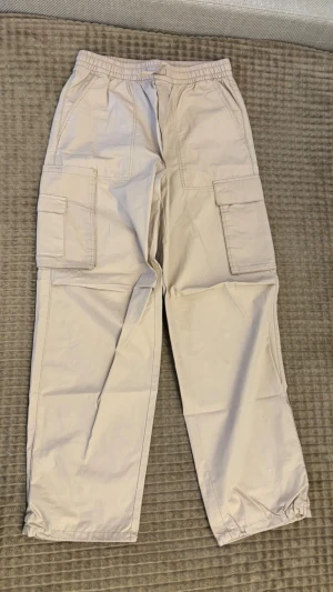 Beige cargobyxor med resårmidja från H&M - Snygga beige cargobyxor med resår i midjan och benslut. Byxorna har stora sidofickor med lock och raka ben. Tillverkade i ett lätt och bekvämt material, perfekta för en avslappnad streetwear-look.