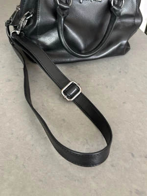 Svart handväska från Fiorelli - Snygg svart handväska från Folli Follie i slätt konstläder. Väskan har både handtag och justerbar axelrem med silverfärgade metalldetaljer och dragkedja upptill. Perfekt för dig som vill ha en stilren och praktisk väska.