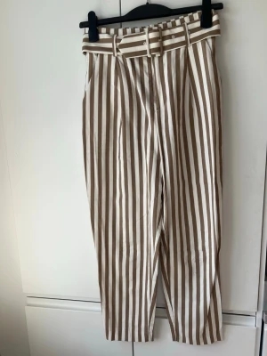 Beigevitrandiga byxor med bälte - Snygga byxor med beige och vita vertikala ränder och matchande bälte i midjan. Byxorna har hög midja och raka ben, vilket ger en trendig och avslappnad look. Materialet känns mjukt och bekvämt. Fläck på knät som säkert går bort med fläckborttagning. 