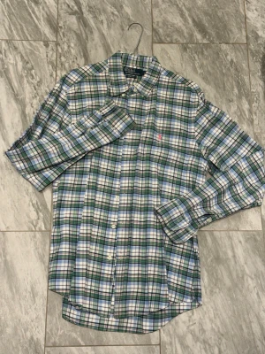 Rutig skjorta från Ralph Lauren - Klassisk rutig skjorta från Ralph Lauren i grönt, blått och vitt. Skjortan har lång ärm, knappar framtill och en liten rosa logga broderad på bröstet. Perfekt för en avslappnad men ändå stilren look.
