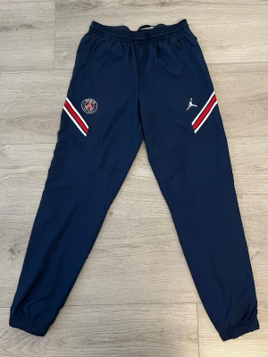 Blå PSG x Jordan träningsbyxor - Mörkblå träningsbyxor från Jordan i samarbete med PSG. Byxorna har elastisk midja, muddar vid benslut och detaljer i rött och vitt på sidorna. PSG-logga och Jumpman-logga framtill. Perfekta för sport eller chill.