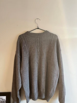 Grå tröja - En stilren grå stickad tröja med kabelstickade detaljer längs framsidan och ärmarna. Tröjan har rund halsringning och ribbstickade muddar vid ärmslut och nederkant. Perfekt för lager-på-lager och en avslappnad look.