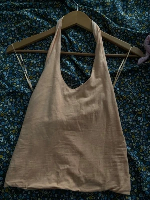 Aprikos linnetopp med halterneck - Snygg beige linnetopp med halterneck och öppen rygg. Toppen har en enkel, stilren design och är tillverkad i ett tunt, mjukt tyg som känns luftigt. Perfekt för varma dagar och enkel att matcha med olika stilar.