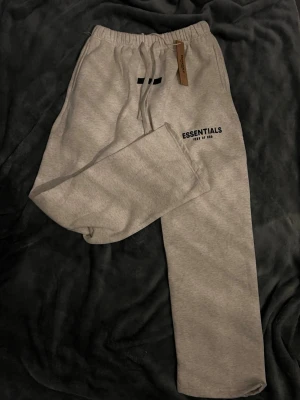 Grå Essentials mjukisbyxor - Snygga grå mjukisbyxor från Essentials Fear of God med svart logga på ena benet. Byxorna har resår i midjan, dragsko och raka ben. Perfekta för en avslappnad och trendig stil. Tillverkade i mjukt material för extra komfort.