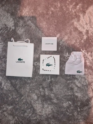 Silverfärgat armband från Lacoste - Snyggt armband från Lacoste i silverfärgad metall med fyrkantiga länkar och diskret Lacoste-logga. Kommer med originalpåse, ask och presentpåse. Perfekt accessoar för dig som gillar stilrena detaljer och vill ha något som sticker ut lite extra. Längd 21cm