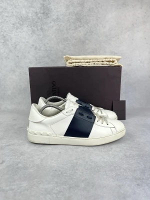 Valentino vita sneakers med svart detalj - Snygga vita sneakers från Valentino med en bred svart läderdetalj över sidan. Skorna har klassisk låg profil, vita snören och subtila nitar på hälen. Tillverkade i slätt läder för en clean och exklusiv look.