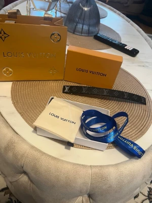 Louis Vuitton svart monogram armband - Snyggt svart armband från Louis Vuitton i läder med klassiskt monogrammönster och silverfärgad LV-logga framtill. Kommer med original ask, påse och blått band. Perfekt accessoar för dig som gillar exklusiv och tidlös design.
