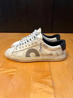 Oliver Cabells vita - Snygga vita sneakers från Oliver Cabell med svart hälparti och grå cirkeldetalj på sidan. Skorna har klassisk låg profil, vita snören och är tillverkade i läder. Defekter syns på bild 4 och 5. Priset är inte hugget i sten