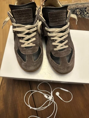 Bruna Maison Margiela Replica sneakers - Säljer ett par Maison Margiela Replica sneakers i brunt mocka och svart läder. Skorna har vita snören, klassisk låg silhuett och ikonisk design med detaljer i både mocka och läder. Perfekta för dig som gillar stilrena och exklusiva sneakers. ÄKTA