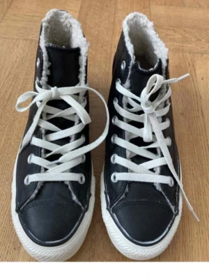 Converse Chuch Taylor All Star, höga speakers i skinn. Stl. US 5 (37,5 svensk) - Klassiska svarta Converse Chuck Taylor All Star med vitt foder i mjuk teddy, vita snören och vit sula. Hög modell med metallhål för snörning och ikonisk logga på sidan. Perfekta för dig som vill ha både stil och värme under kallare dagar. Innermått 23,2 cm