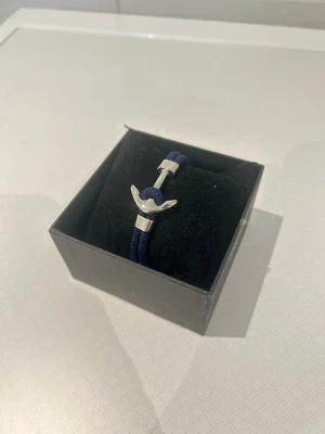 Marint armband med ankardetalj - Snyggt armband med marinblått rep och en silverfärgad ankare som lås. Perfekt accessoar för dig som gillar nautisk stil. Armbandet har en enkel men stilren design och passar till många olika outfits.