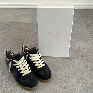 Maison Margiela Replica svarta sneakers - Säljer ett par svarta sneakers från Maison Margiela. Väldigt fint skick och väl omhändertagna. Köpta från Maison Margielas officiella hemsida. Nypris: 6 600 kr / 610 €. Allt medföljer: box, dustbag, QR-kod och övriga tillbehör se bild 4. Äkthetsbevis kan skickas vid önskemål. Säljes pga kommer tyvärr inte till användning. Stilrena och tidlösa – perfekta för dig som gillar minimalistisk design med streetwear-känsla.
