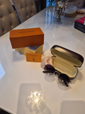 Louis Vuitton solglasögon guld/svart - Snygga solglasögon från Louis Vuitton med guldfärgade metallbågar och svarta, tonade glas. Runda linser och exklusiv känsla, levereras med originalfodral, putsduk och box. Perfekt accessoar för dig som vill sticka ut med stil.