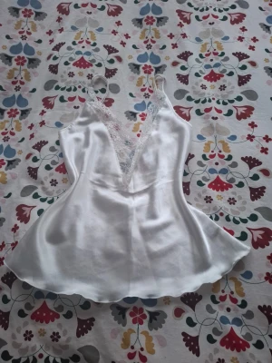 Vit satinlinnetopp med spets - Snygg vit linnetopp i satin med djup v-ringning och spetsdetaljer vid bysten. Tunt axelbandslinne med lätt glansig finish och feminin siluett. Perfekt att styla med jeans eller kjol för en trendig look.