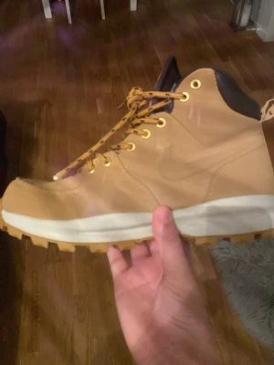 Nike ljusbruna boots med snörning för vintern  - Säljer ett par ljusbruna boots från Nike med grov sula och snörning i guldiga öljetter. Skorna har svart vadderad kant upptill och är tillverkade i slitstarkt läder. Perfekta för dig som vill ha en streetig och robust look.