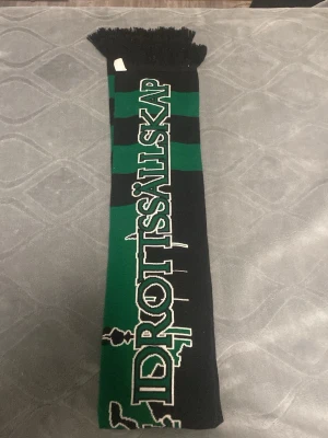 GAIS grönsvart halsduk med text - Snygg halsduk i grönt och svart med texten 'Göteborgs Atlet & Idrottssällskap'. Perfekt accessoar för dig som vill visa ditt GAIS-stöd. Halsduken har fransar i ändarna och är stickad i mjukt material.