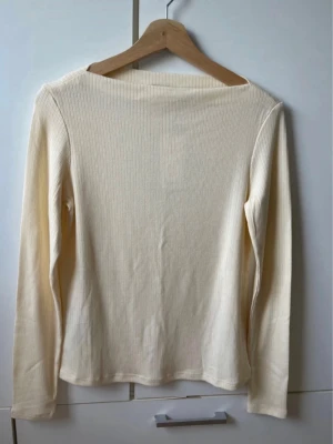 Ljusribbad långärmad topp - En enkel och stilren cremevit/ljusgul långärmad topp med ribbad struktur. Toppen har en rak passform och är tillverkad i ett mjukt material som känns skönt mot huden. Perfekt att matcha med jeans eller kjol för en clean look.