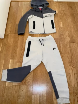 Nike grå och vit tracksuit set - Säljer ett snyggt tracksuit set från Nike i grått och vitt. Setet består av en hoodie med dragkedja och två svarta fickor samt matchande byxor med svarta detaljer och dragsko i midjan. Perfekt för träning eller chill. Materialet är mjukt och stretchigt.