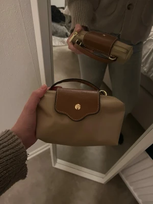 Beige handväska  - Snygg beige handväska. med bruna läderdetaljer och guldfärgad knapp. Väskan har ett justerbart axelband i brunt läder och en praktisk dragkedja. Perfekt storlek för det viktigaste och enkel att bära crossbody.