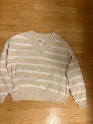 Beige och vit randig stickad tröja - Mysig stickad tröja från hollister i beige och vitt med breda horisontella ränder. Tröjan har V-ringning, långa ärmar och ribbade muddar vid ärmslut och nederkant. Perfekt för lager på lager och chill stil. Använd en gång. Storlek xs. Frakt ingår ej!