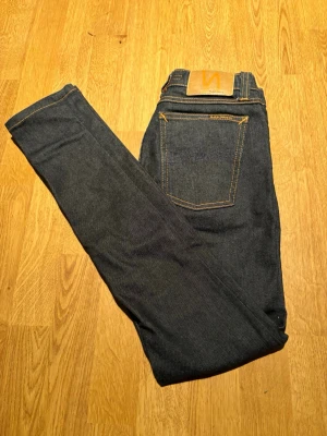 Mörkblå jeans från Nudie Jeans - Snygga mörkblå jeans från Nudie Jeans med klassisk femficksdesign och kontrastsömmar. Jeansen har rak passform och är tillverkade i kraftig denim med läderpatch bak i midjan. Perfekta för en stilren och avslappnad look.