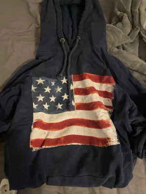 Mörkblå hoodie med USA-flagga - Säljer en mörkblå hoodie med stor amerikansk flagga tryckt på bröstet. Hoodien har huva med dragsko och en avslappnad passform. Perfekt för dig som gillar streetwear och vill sticka ut med en bold print.