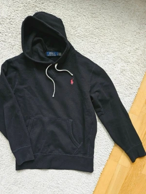 Svart hoodie från Polo Ralph Lauren - Snygg svart hoodie från Polo Ralph Lauren med klassisk huva och vita dragsnören. I helt nytt skick använts fåtal gånger. Har den ikoniska röda loggan broderad på bröstet och en stor magficka framtill. Perfekt för en chill och stilren look. Materialet är mjukt och skönt bomullstyg.
