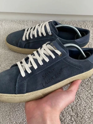  Saint Laurent - Mörkblå sneakers i mocka från Saint Laurent med broderad logga på sidan. Skorna har vita snören, beige sula och klassisk låg profil. Perfekta för dig som gillar stilrena och exklusiva sneakers med en lyxig känsla. Skick 8-9/10 fruktigt schyssta skor 