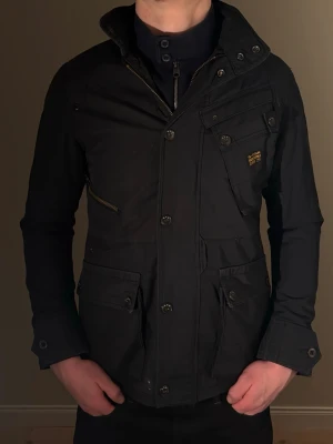 Mörkblå G-Star RAW jacka - Snygg mörkblå jacka från G-Star RAW med flera fickor, dragkedja och knappar framtill. Jackan har hög krage och detaljerade sömmar. Storlek m men är något tightare i modellen. Jackan är i mycket bra skick, har däremot en liten blekt fläck på ryggen, ej märkbar i helhet.