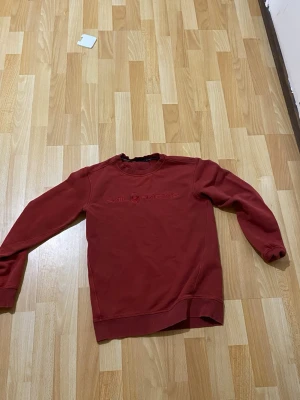 Röd crewneck från Sail Racing - Säljer en snygg röd crewneck från Sail Racing med broderad logga på bröstet. Tröjan har rund hals, ribbade muddar och är tillverkad i mjukt bomullsmaterial. Perfekt för en avslappnad och stilren look.