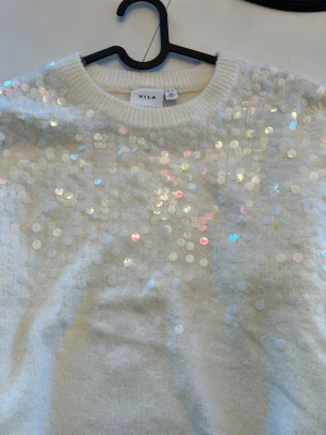 Vit paljettstickad tröja från Vila - Superfin vit stickad tröja från Vila med massor av skimrande paljetter framtill. Tröjan har rund halsringning och är perfekt för dig som vill sticka ut lite extra. Mjuk och bekväm, passar perfekt till jeans eller kjol.