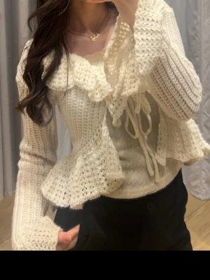 stickad volangkofta - Supersöt krämvit stickad kofta med volangdetaljer och knytband framtill. Koftan har långa ärmar med volang vid ärmslut. Perfekt att styla över ett linne eller klänning. Materialet är mjukt och luftigt.