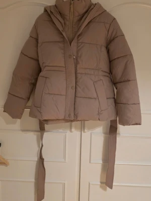 Beige pufferjacka med huva och bälte - Snygg beige pufferjacka med huva och breda kanaler. Jackan har ett matchande bälte i midjan och stora fickor framtill. Stängs med dragkedja och tryckknappar. Aldrig använd. Står storlek M men passar mer åt en S