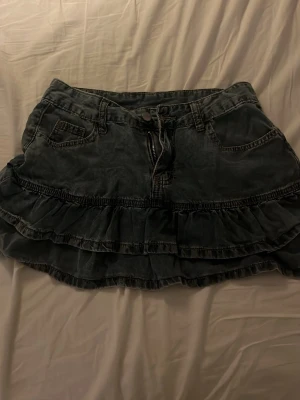 Kort jeanskjol med volanger - Säljer en mörkblå jeanskjol med volangdetaljer och fickor fram. Kjolen har en klassisk jeanslook med knapp och dragkedja framtill samt två lager volanger som ger en cool och trendig vibe.