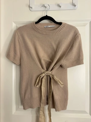 Beige stickad topp med knytband Zara - Snygg beige stickad topp från Zara med korta ärmar och ribbade muddar. Toppen har ett dekorativt knytband framtill som ger en trendig look. Perfekt till jeans eller kjol för en avslappnad stil.