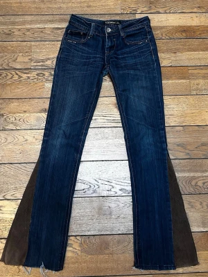 Blå utsvängda jeans från Redpepper - Snygga mörkblå jeans från Redpepper med unika bruna kilar i manchestertyg längs bensluten. Jeansen har fem fickor, låg midja och råa kanter nertill. Perfekt för dig som vill sticka ut med en cool och retroinspirerad look. Utsydda av mig