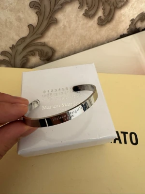 Maison Margiela silverarmband - Snyggt och stilrent armband från Maison Margiela . Armbandet är öppet och har en minimalistisk design med ingraverad logotyp och serienummer. Perfekt accessoar för dig som gillar exklusiva detaljer och modern stil.                        Oops detta är 1:1 i rostfritt stål så mer eller mindre identiskt 