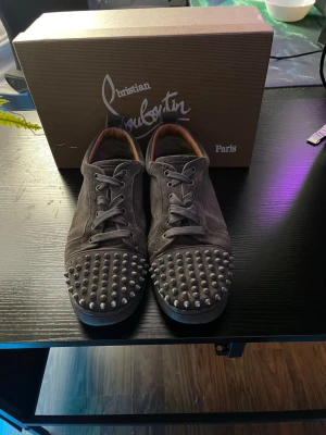  Christian Louboutin  - Unika gråa sneakers från Christian Louboutin i mocka med coola silverfärgade nitar på tån. Tre nitar saknas och färg har försvunnit.  Lågt skaft och klassisk snörning. Insidan är fodrad i brunt läder för extra komfort. Perfekta för dig som vill sticka ut med din stil.