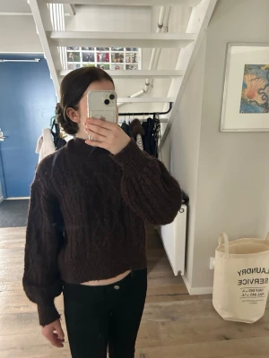 Brun kabelstickad tröja från KappAhl - Mysig brun kabelstickad tröja med hög hals och ballongärmar. Tröjan har ett kortare, boxigt snitt och breda ribbade muddar vid ärmslut och nederkant. Perfekt för lager-på-lager och höstiga outfits.