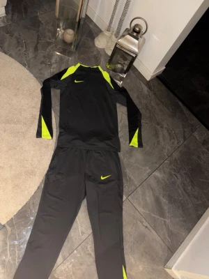 Nike svart träningsset med neon - Säljer ett svart träningsset från Nike med långa ärmar och byxor. Setet har snygga neon-gula detaljer på axlar, ärmar och ben samt Nike-logga på både tröja och byxor. Tillverkat i ett lätt och stretchigt funktionsmaterial som passar perfekt till träning.