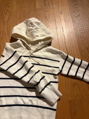 Vit hoodie med marinblå ränder - Snygg vit hoodie med marinblå horisontella ränder och dragsko i huvan. Tröjan har långa ärmar och är tillverkad i ett mjukt stickat material som känns skönt mot huden. Perfekt för en avslappnad och stilren look.