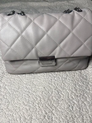 Grå handväska med kedjerem - Snygg quiltad handväska i ljusgrått med silverfärgad kedjerem och metallspänne. Väskan har en klassisk rektangulär form och är tillverkad i syntetmaterial med mjuk känsla. Perfekt accessoar för att lyfta din outfit.
