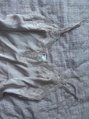 Ljusrosa satinpyjamas med spetsdetaljer - Säljer ett tvådelat pyjamasset i ljuslila satin med tunna axelband och vacker spets längs urringning och benslut. Setet består av ett linne och shorts, båda med mjuk och glansig finish. Perfekt för mysiga kvällar hemma.
