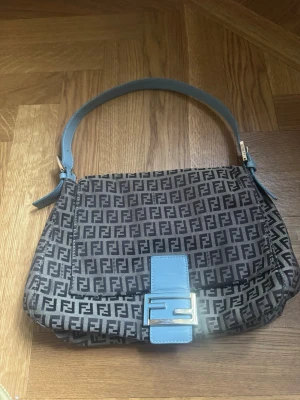 Fendi Baguette monogram handväska blå - Säljer en ikonisk Fendi Baguette handväska med klassiskt FF-monogram i svart och brun canvas tyg. Väskan har ljusblått läderhandtag och matchande blått spänne med metallogo framtill. Perfekt axelväska för dig som gillar statement-plagg och designerstil.